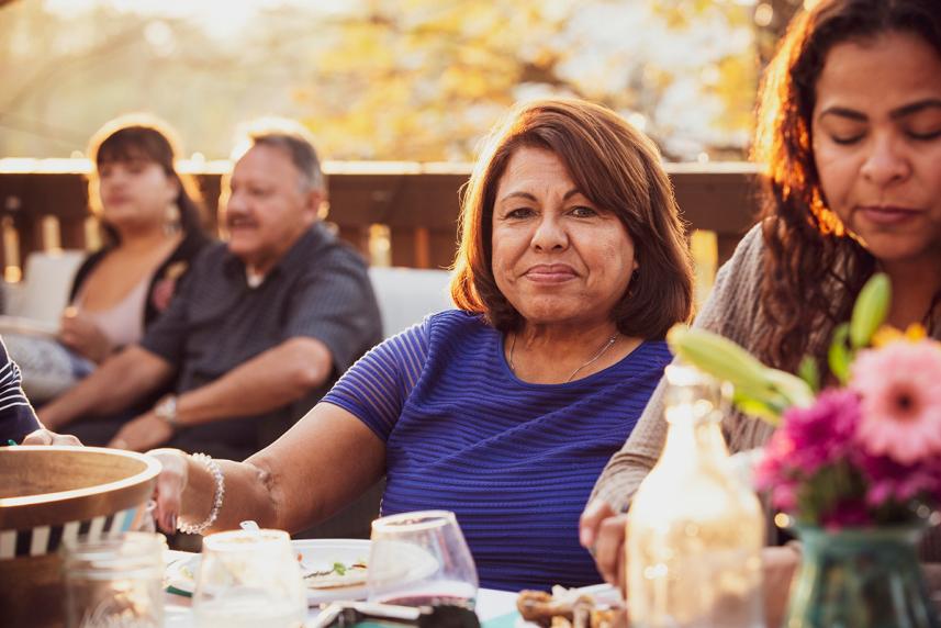 A group of people sitting outside at a table with food, drinks, and flowers - Woman staring at camera